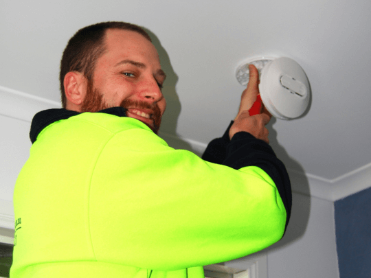 Bearded man wearing yellow hi-vis jumper installing a white smoke alarm.